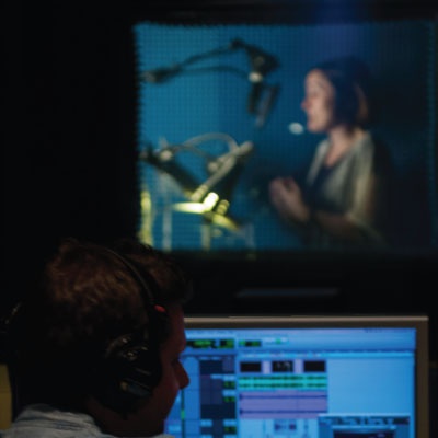 Spanish voice talent reading script in booth, while studio engineer monitors levels in the foreground.